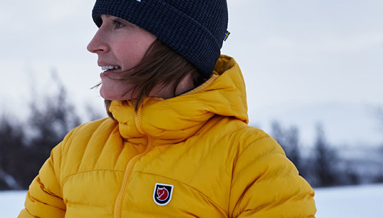 En kvinde i gul Fjällräven dunjakke i et snelandskab.