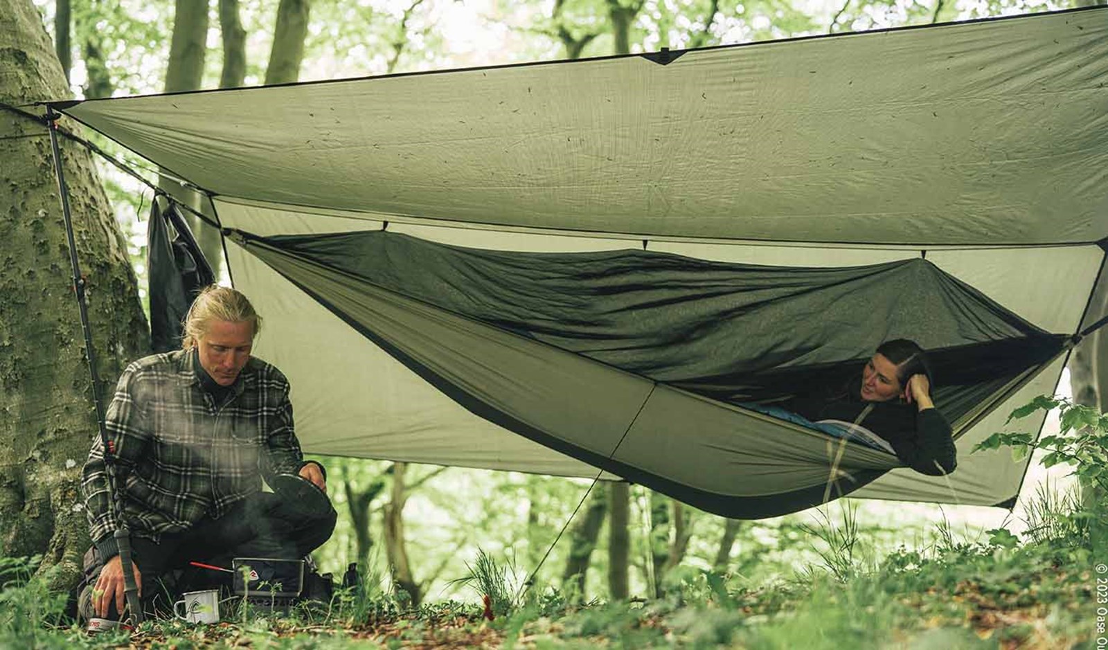 To personer under en tarp i skoven, hvoraf den ene ligger i en hængekøje.