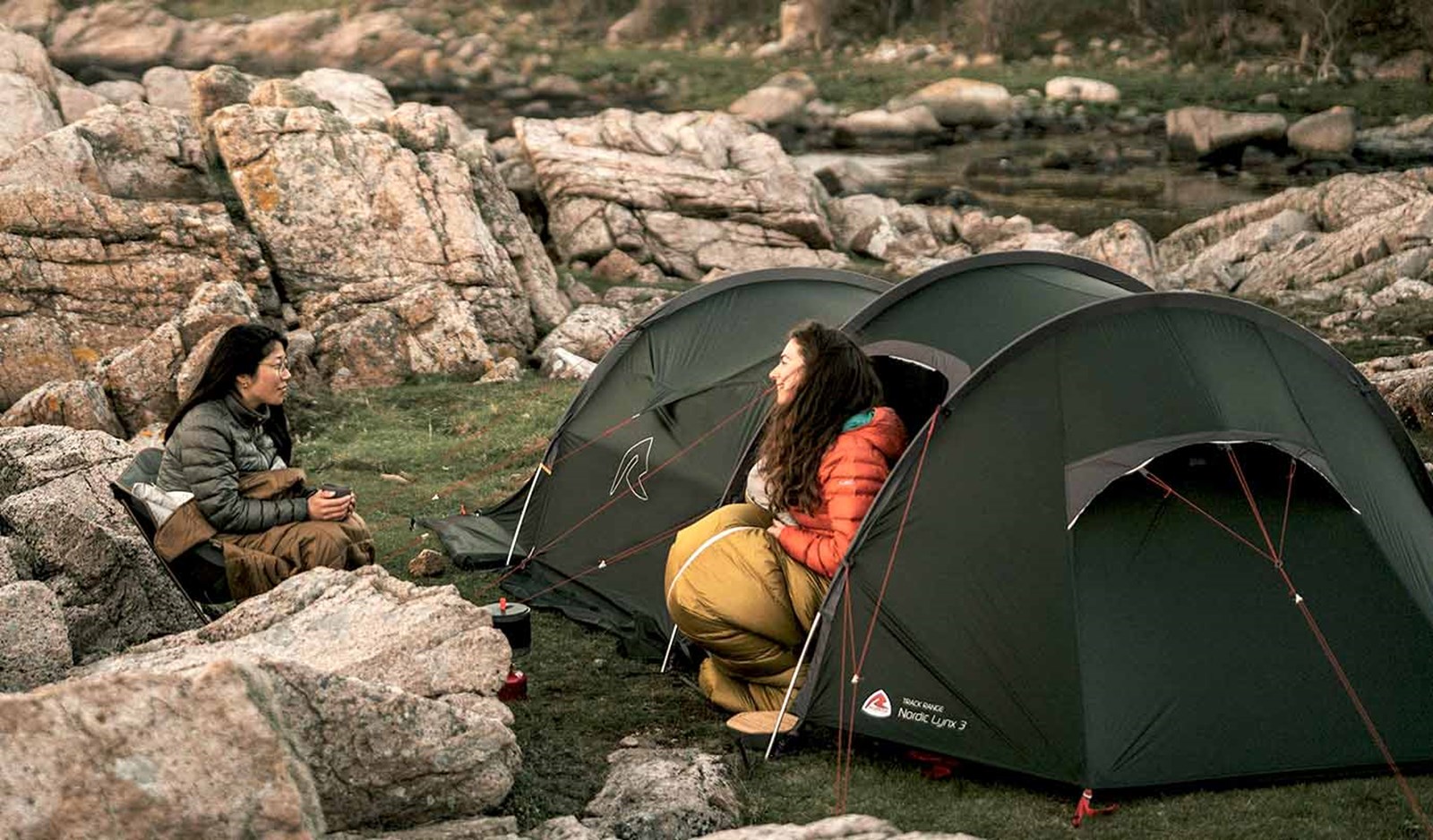 To personer sidder og snakker iført soveposer ved deres tunneltelt.