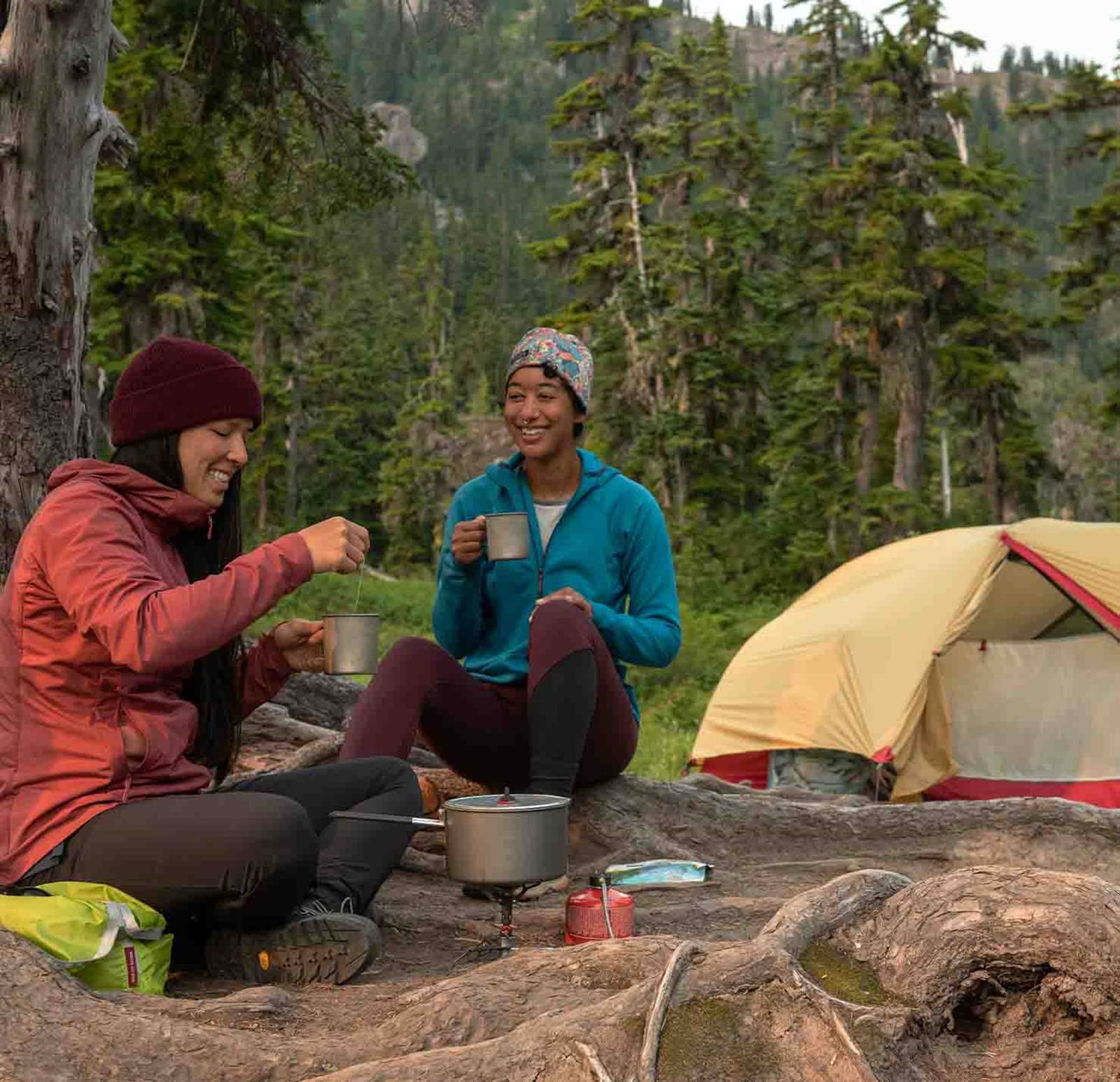To personer sidder og nyder en kop te i naturen, med et telt i baggrunden.