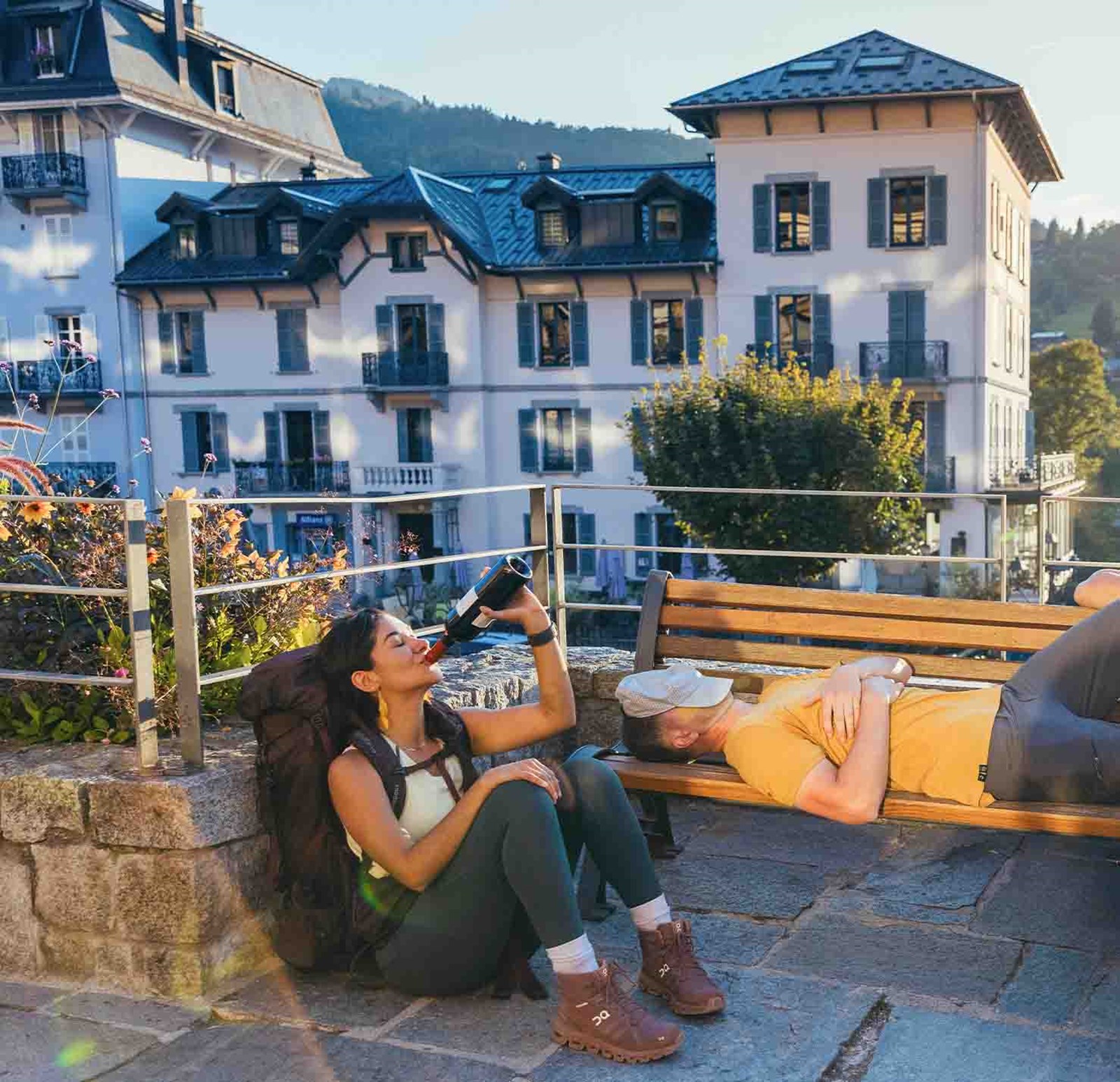 En kvindelig backpacker bruger ventetiden på at drikke vin, mens hendes makker tager en lur på en bænk.