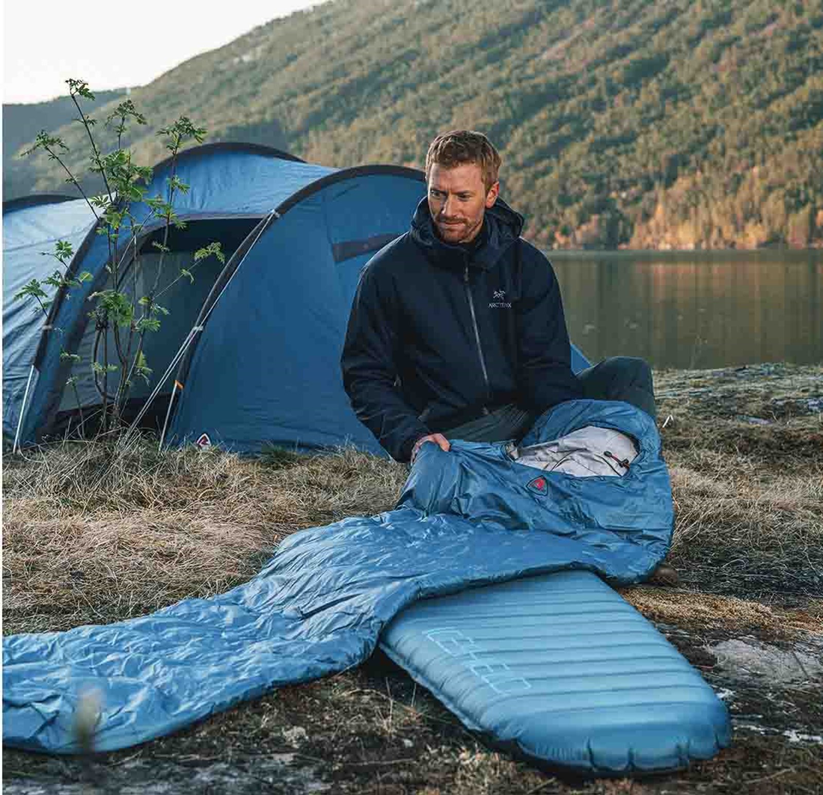 En mand der sidder med sit sovesetup ude i naturen foran hans telt.