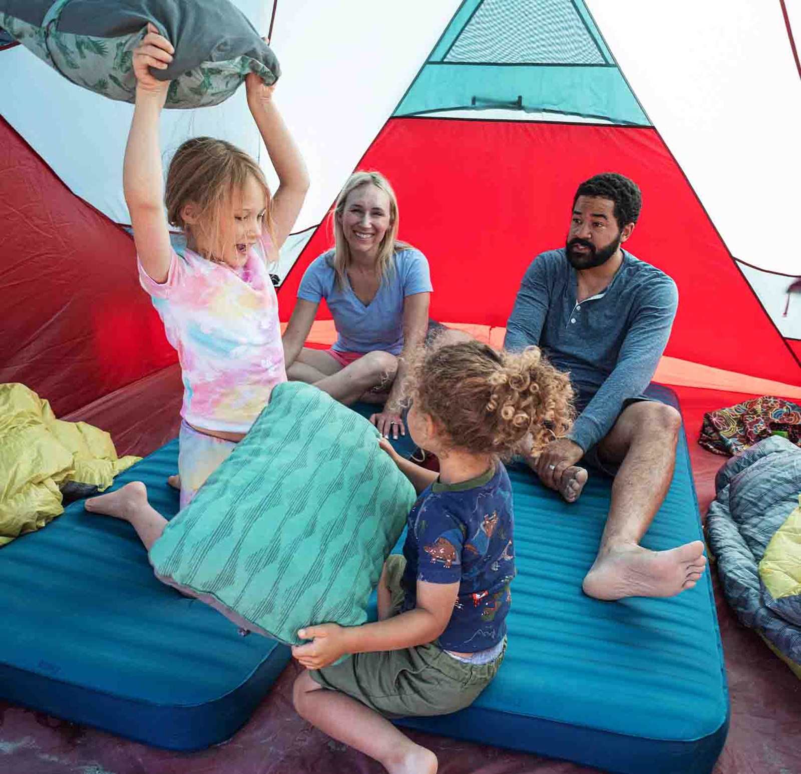 En familie sidder i et telt og leger pudekamp på deres lidt større selvoppustende liggeunderlag.