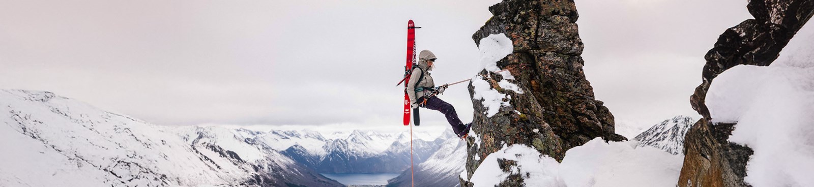 Person der klatrer på klippe med ski på ryggen i en skaljakke.